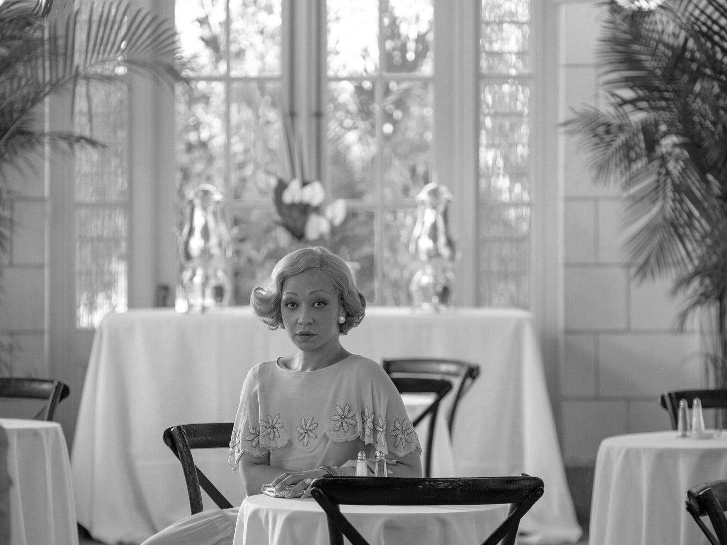 Black & White photo of Ruth Negga with blonde hair and a white dress in a tea room with white curtains and table cloths.