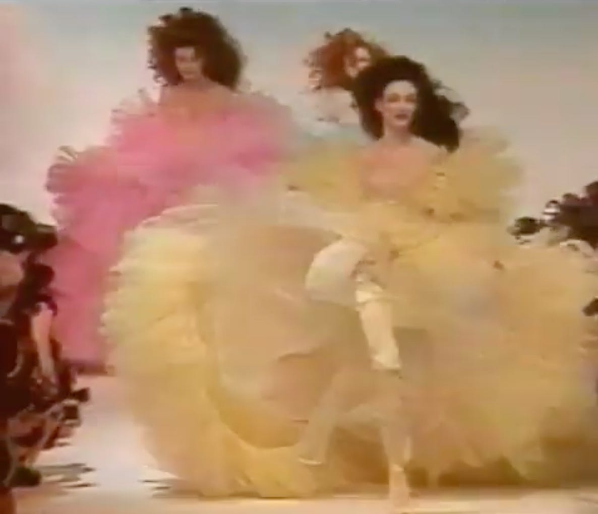 A blurry Thierry Mugler catwalk photo, three white figures with large curly hairdos wear bouffant fluffy dresses in pastel shades. 