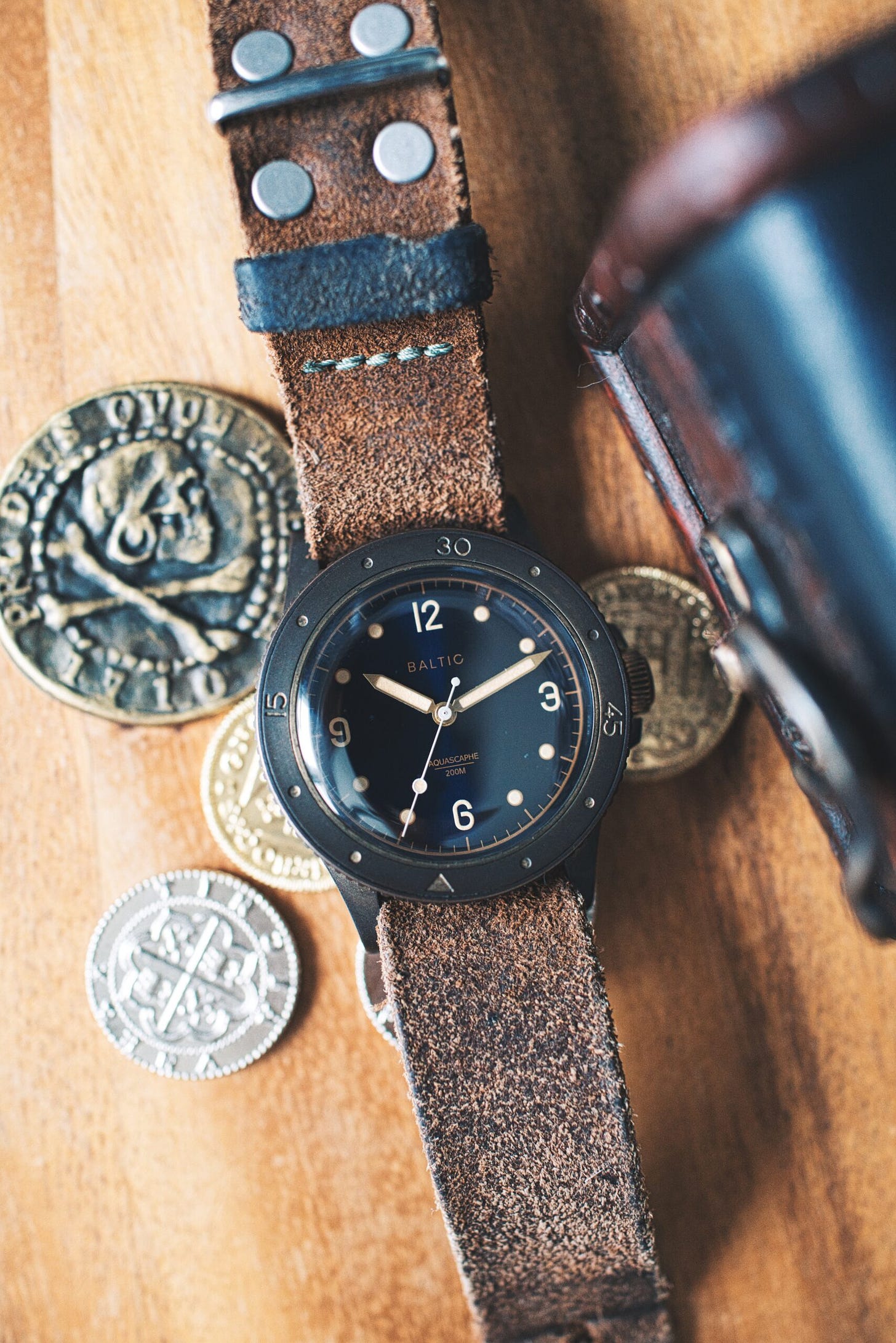 A very patinaed baltic bronze next to old pirate coins, near a small treasure chest sitting atop a wood desktop. Watch on a worn rough leather strap.