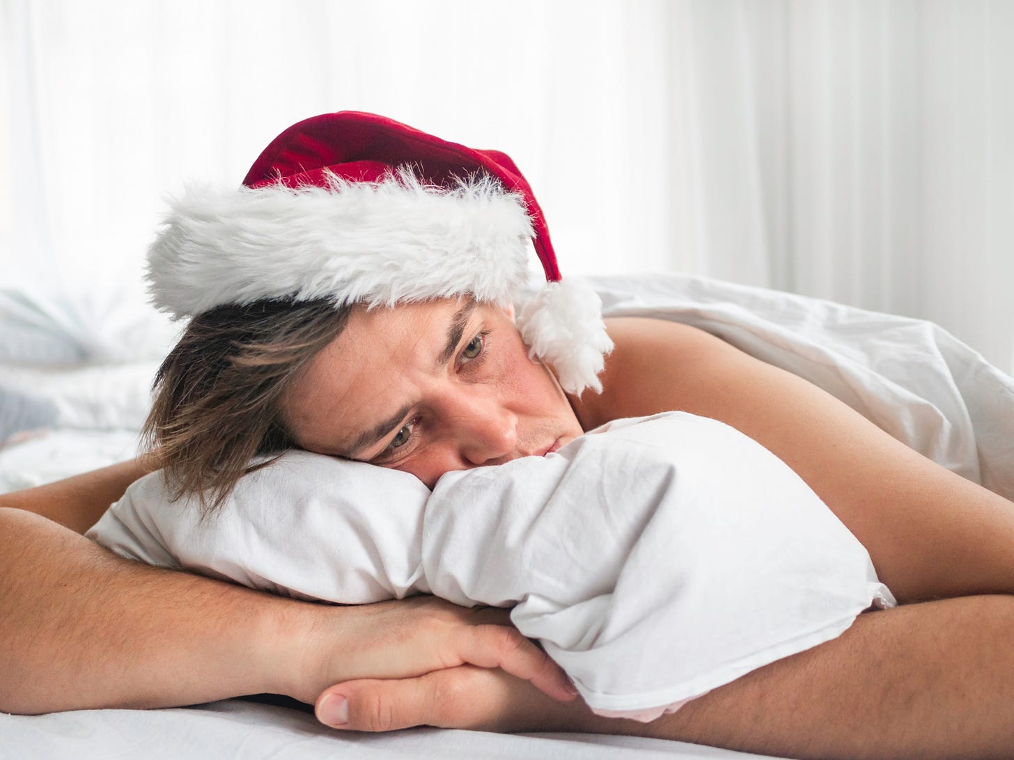 A sad man in a santa hat hugs his pillow and stares into the distance.