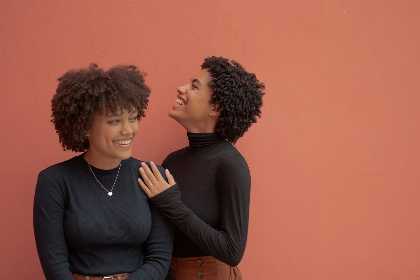 Two Black women smile and laugh together. They both wear black turtlenecks.