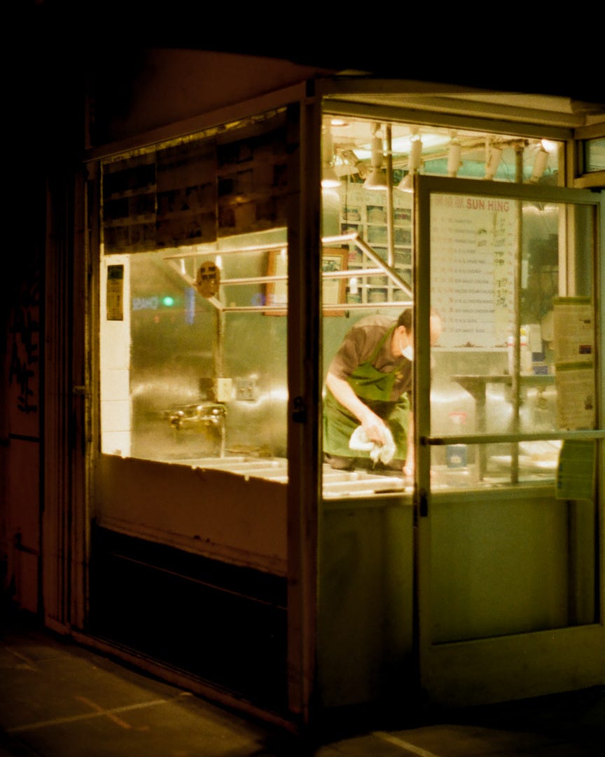 A restaurant worker cleaning up in a storefront window. Inside, the shop is yellow light and the rest of the environment is dark.