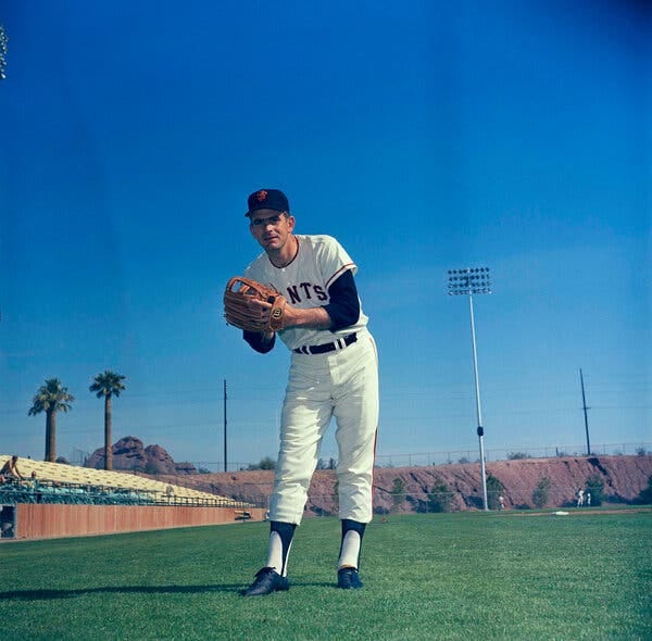 A color photo of Perry in a white Giants uniform and black cap in a pitcher’s pose on a carpet of green grass. A couple of palm trees are in the distance.