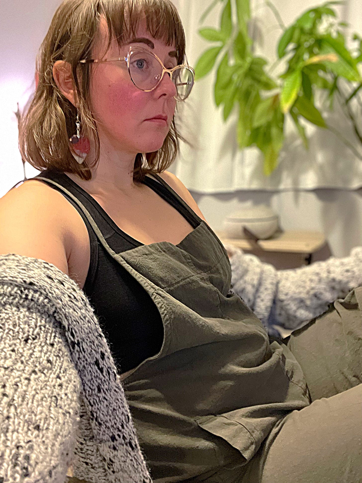 Dr. bird in gold cat eye glasses, red and white tear drop acrylic earrings, a black sports bra, green overalls, and a white cardigan with black speckles.