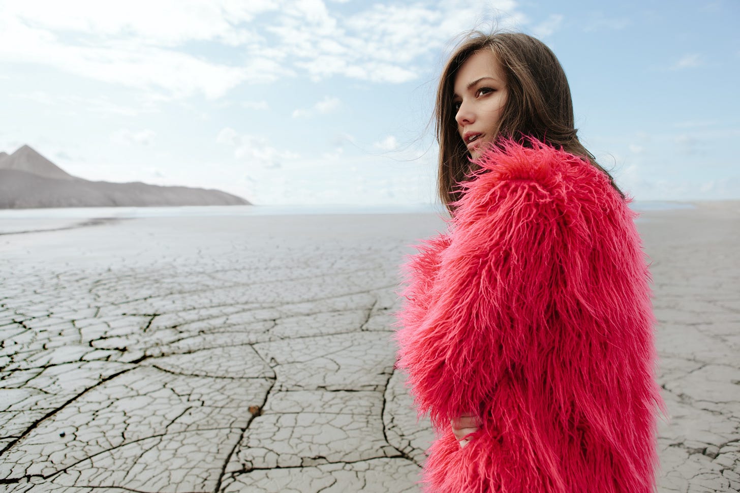 A woman in a shaggy pink coat stands against a desert backdrop.