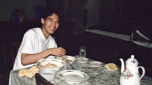 Jeff sitting at a dining table covered with plastic floral tablecloth, eating fufu and light soup with one hand
