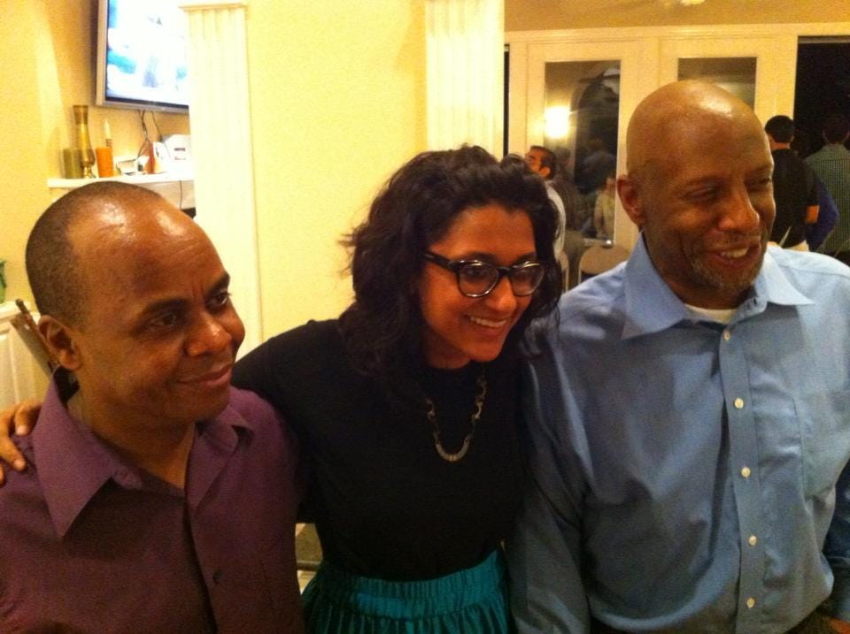 A photo of three people: Two men with a woman in the middle, with arms around both to pose. The men are both people of color, and bald, and Deepti, at center, is a mid-30s Indian woman with glasses, a black top and a teal skirt and glasses. The men wear button-down shirts, all smiles.