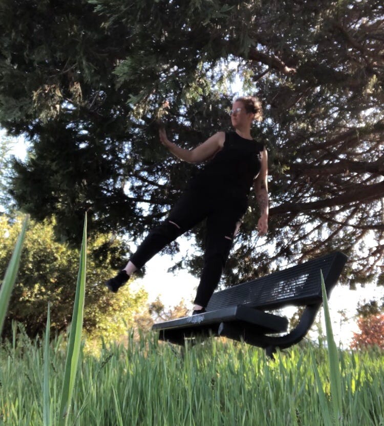 a human dressed all in black mid-dance move on a bench framed by grass and a large tree