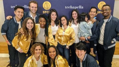 a mixed group of men and women in gold and navy blue jackets smiling in front of a backdrop that says techapella