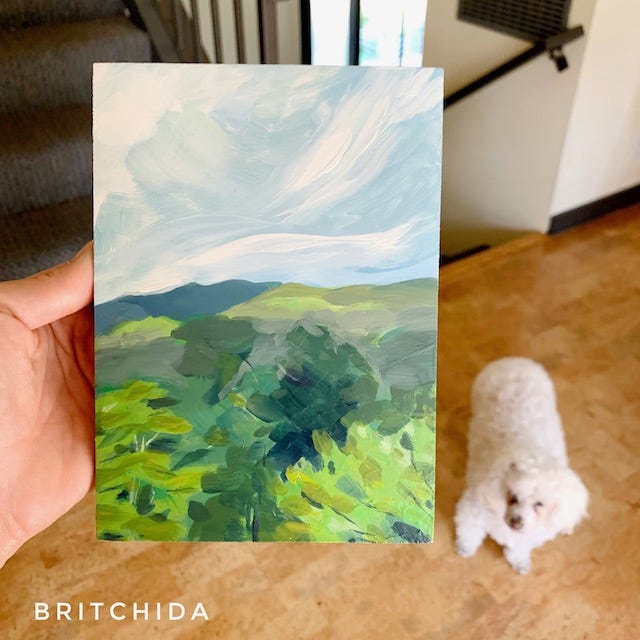 a small landscape painting in blues and greens held up, Elsa the dog in the background. The second image is the same piece with a smaller similar one, sitting on my desk.