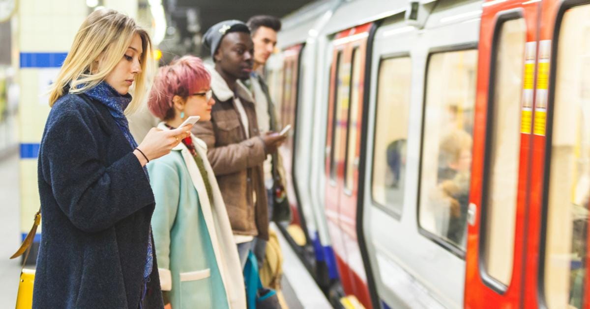 4 commuters in London are waiting on a platform. A London Underground train is in front of them. 2 people are using their smartphones