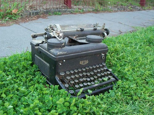 An old Royal typewriter sits in some grass next to a sidewalk. "Royal Typewriter" by avlxyz is licensed under CC BY-SA 2.0 