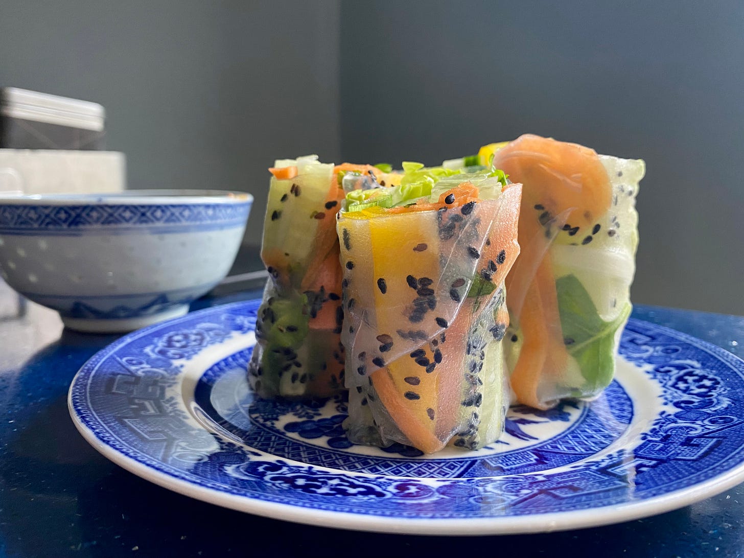 Rice paper rolls arranged on a blue and white plate. Through the rice paper wrappers, carrot, peppers, lettuce and black sesame seeds are visible