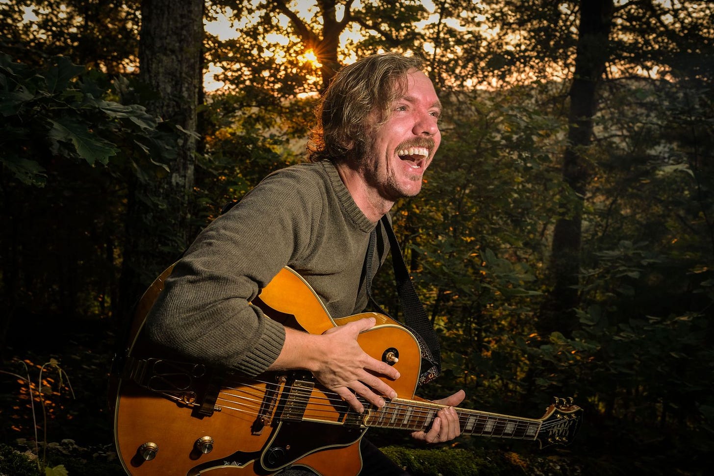 A blonde man cradling an acoustic guitar stands in a forest background as the sun sets through the trees behind him. He is laughing and looking away from the camera. His smile is beautiful. It's Michael De Backer.