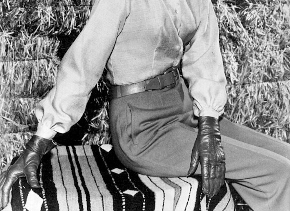 A black and white photo of a woman seated on a blanket in front of bales of hay. She has on pants cinched with a wide leather belt and leather gloves. She can only be seen from just below her shoulders to her knees but we know from the essay that this is Barbara Stanwyck in her role as the matriarch of the TV show "Big Valley." 