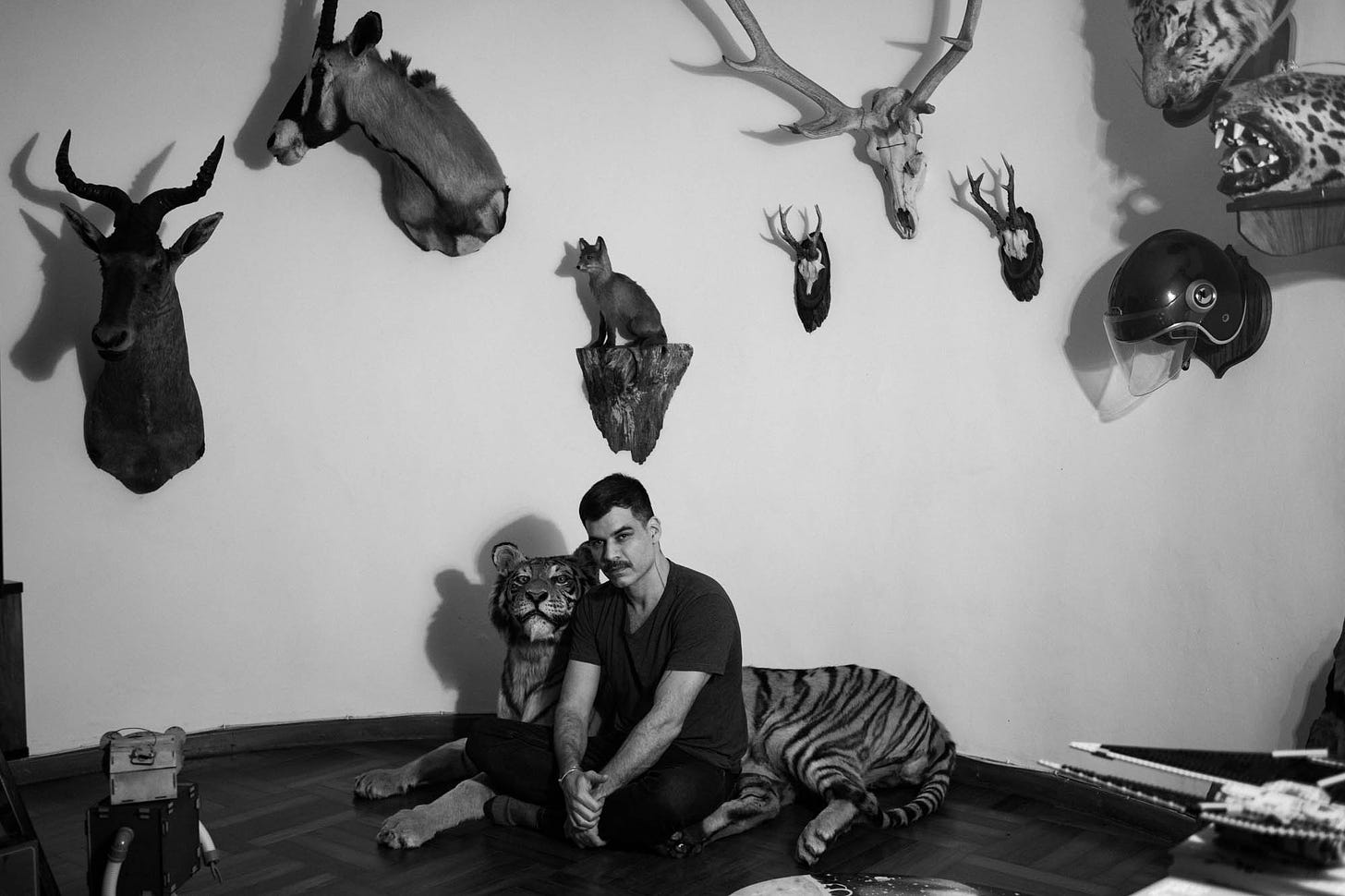 iamgem em preto e branco mostra homem sentado numa sala cercado de animais espalhados como uma onça e uma raposa, ele vesta ecalça e camiseta preta e olha diretamente para a câmera.