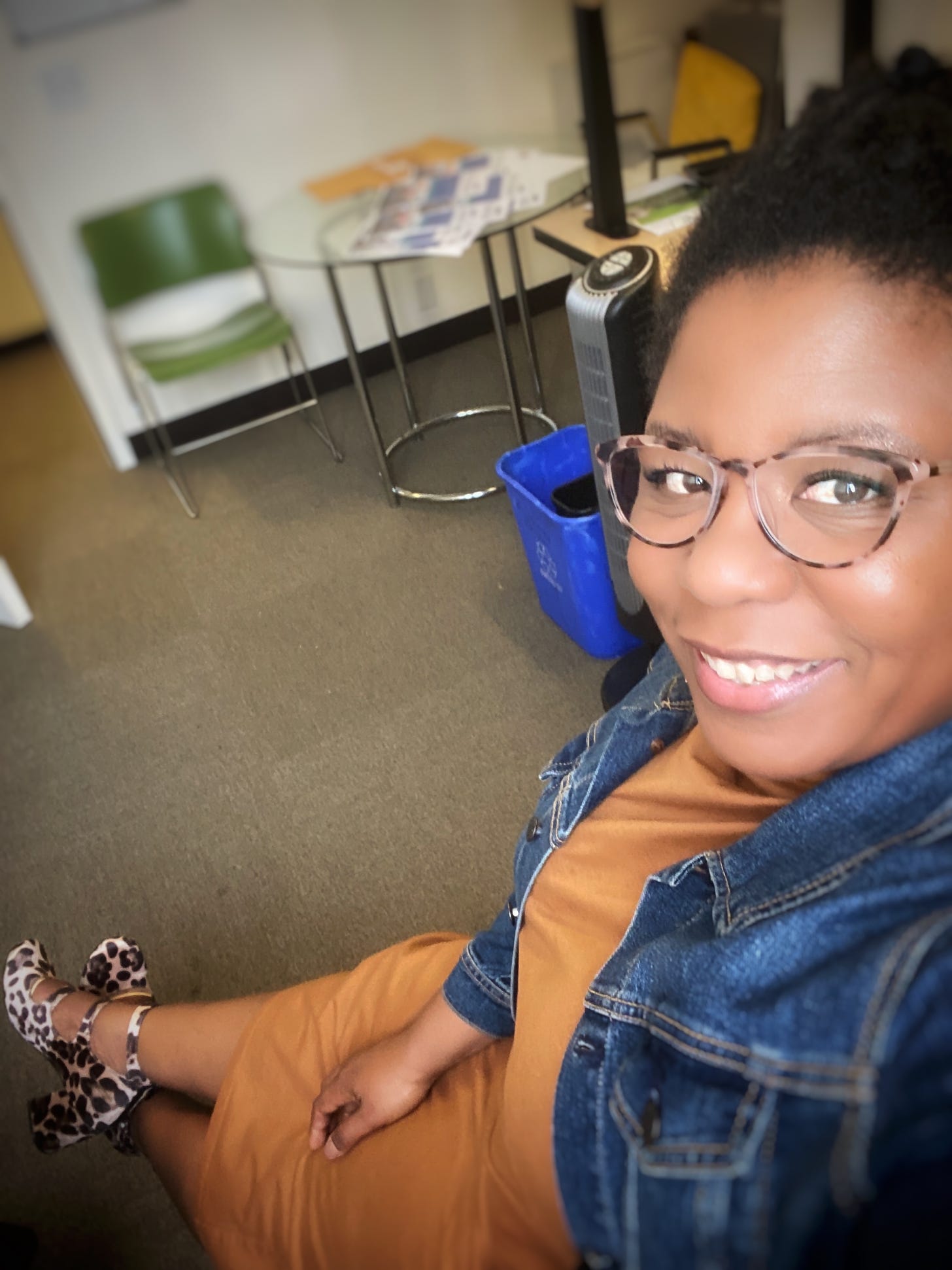 Qianya sits at a desk with her legs in front of her, smiling up towards the camera.  She is wearing a mustard dress with a blue denim jacket.  She has on kick ass cool leopard print block heel shoes and cat eyes glasses.