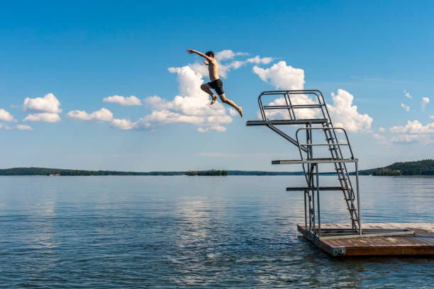 Side view of a teenage male jump diving from a diving tower with blue sky and horizon in the background. Side view of a teenage male jump diving from a diving tower with blue sky and horizon in the background. Hot summer afternoon at a bath place in lake Mälaren in Sweden, horizontal composition. diving platform stock pictures, royalty-free photos & images