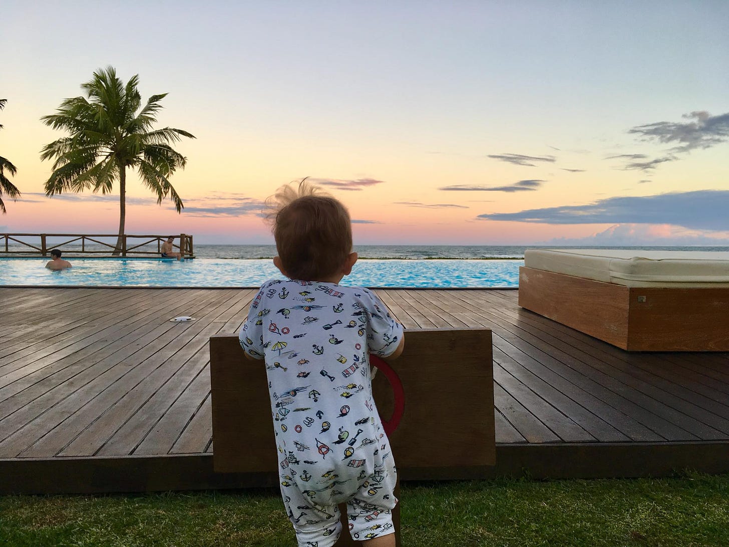 foto do domenico, de costas, apoiado em um deck de madeira, olhando em direção ao mar com um belo céu de fim de tarde na bahia