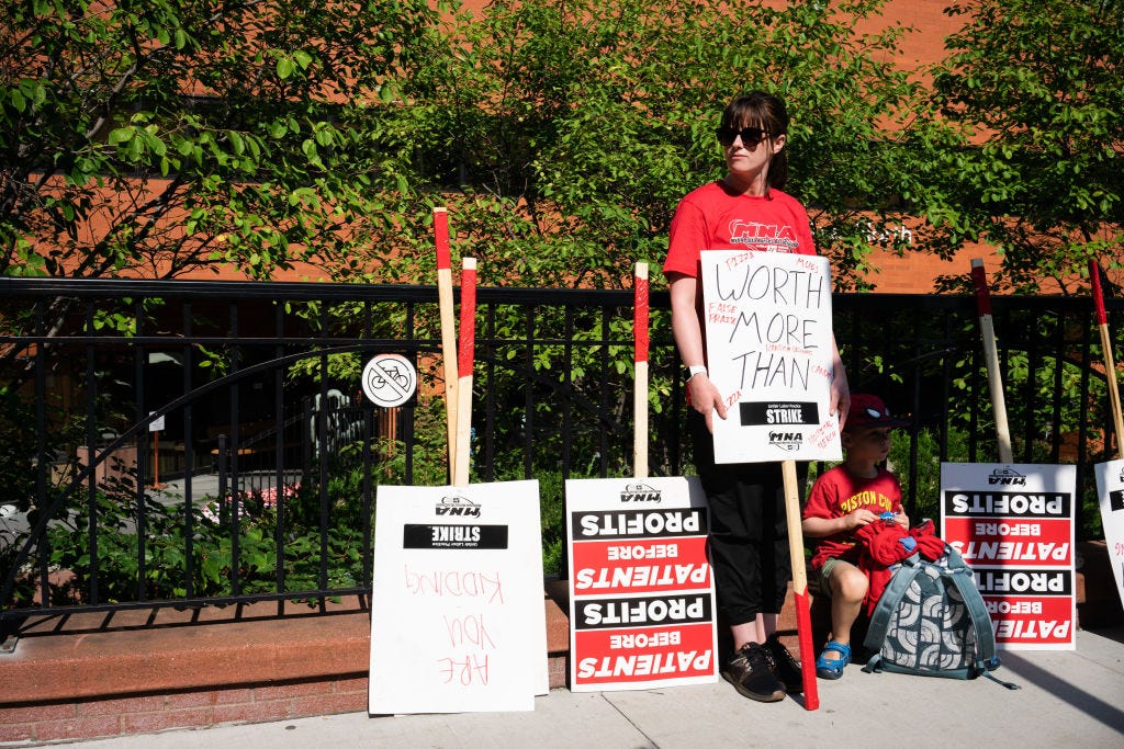 a brunette woman stands against a black iron fence and green trees holding a picket sign leaning against the ground that reads "worth more than" and listing words. other picket signs are leaning against the iron fence and a child is sitting below the woman