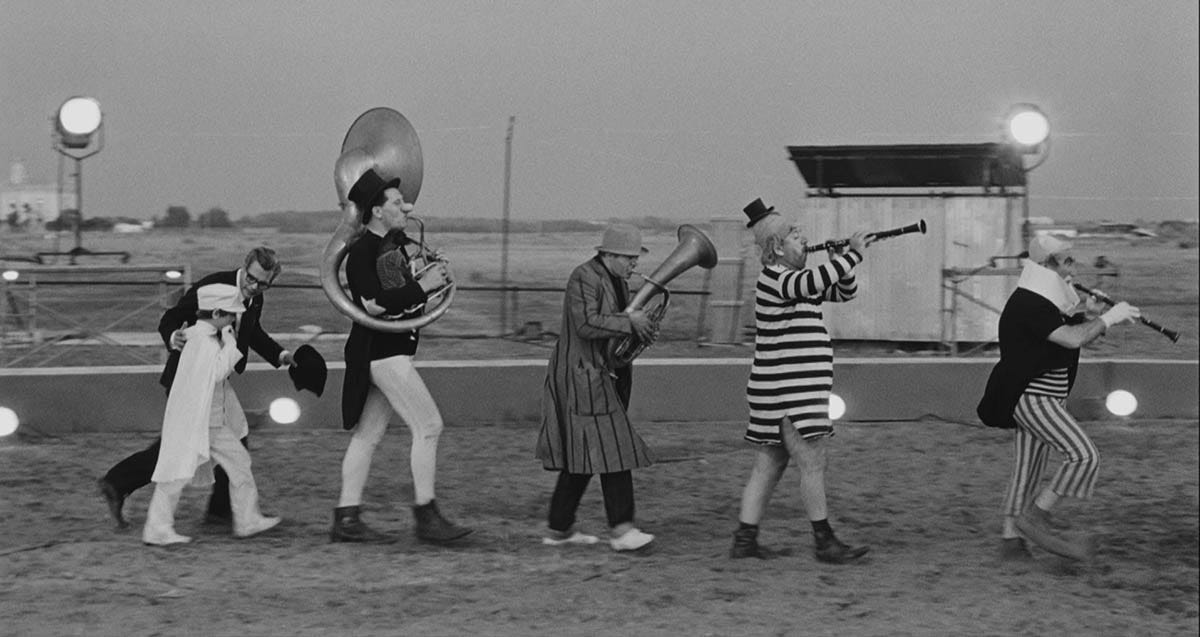 Image: A still from 8 1/2, where Guido leads a child ring leader and several clown musicians around a ring