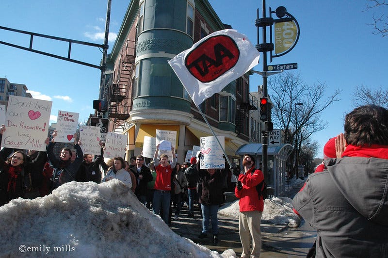 A group of students hold protest signs and wave flags as they stand in the middle of a snowy intersection, the buildings of State Street behind them