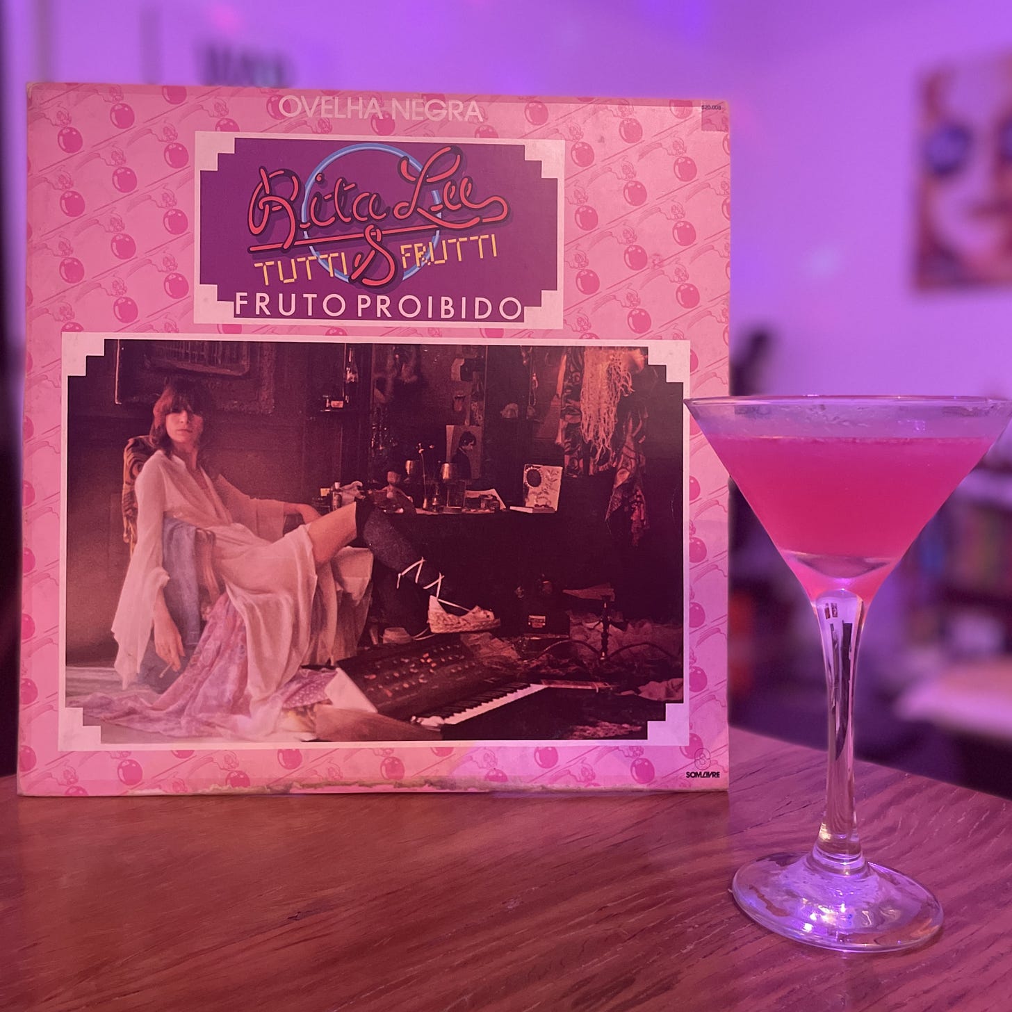 Foto mostra a capa do disco Fruto Proibido, de Rita Lee, em tons rosas, com uma foto da cantora sentada com roupas diáfanas, e uma taça de Martini com um líquido rosa dentro -- é um drink Cosmopolitan