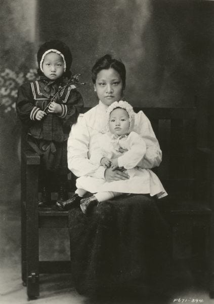 Little Anna dressed in a white dress and bonnet sits in her mother’s lap, while her older sister Lulu stands next to them on the bench, circa 1905