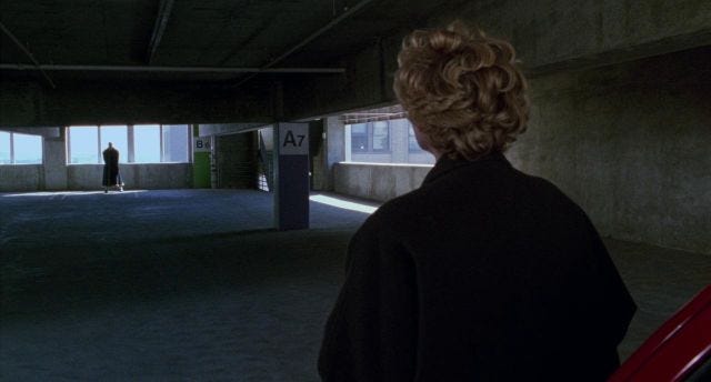 Over the shoulder shot of Virginia Madsen in a parking garage. At the other end of the space is a man in silhouette.