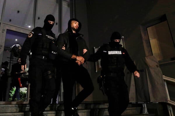 Two uniformed officers escorting Andrew Tate, who is dressed in a black hooded sweatshirt and pants, down the stairs outside the headquarters of Romania’s organized crime agency.