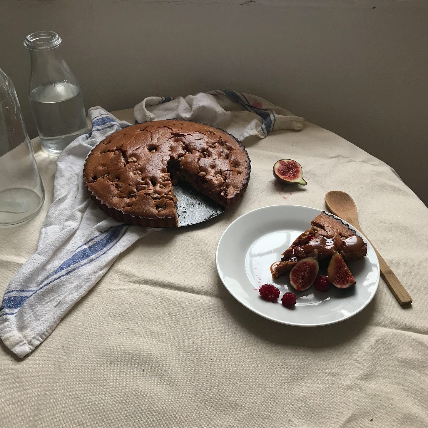 A brown cake still in its tin, on a napkin on a table. The cake has dimples where soft raspberries have cooked into its top. A big segment has been sliced out and arranged on a white plate beside, some cut figs on the plate, one half open on the table. A wooden spoon, and two carafe of water are arranged beside. 