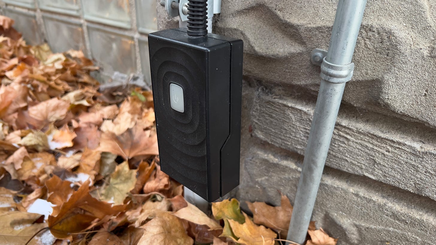 Meross outdoor smart plug hangs by a stone wall over some leaves