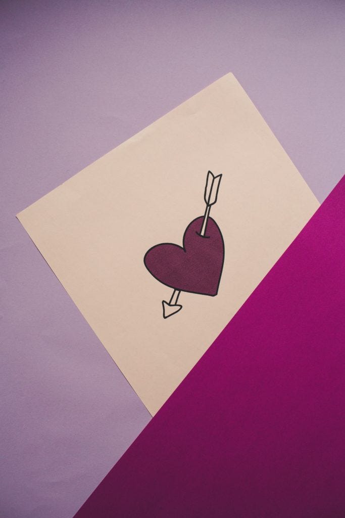 A dark purple heart with an arrow going through it is drawn on a white piece of paper. The background is light purple with a magenta bottom right corner.
