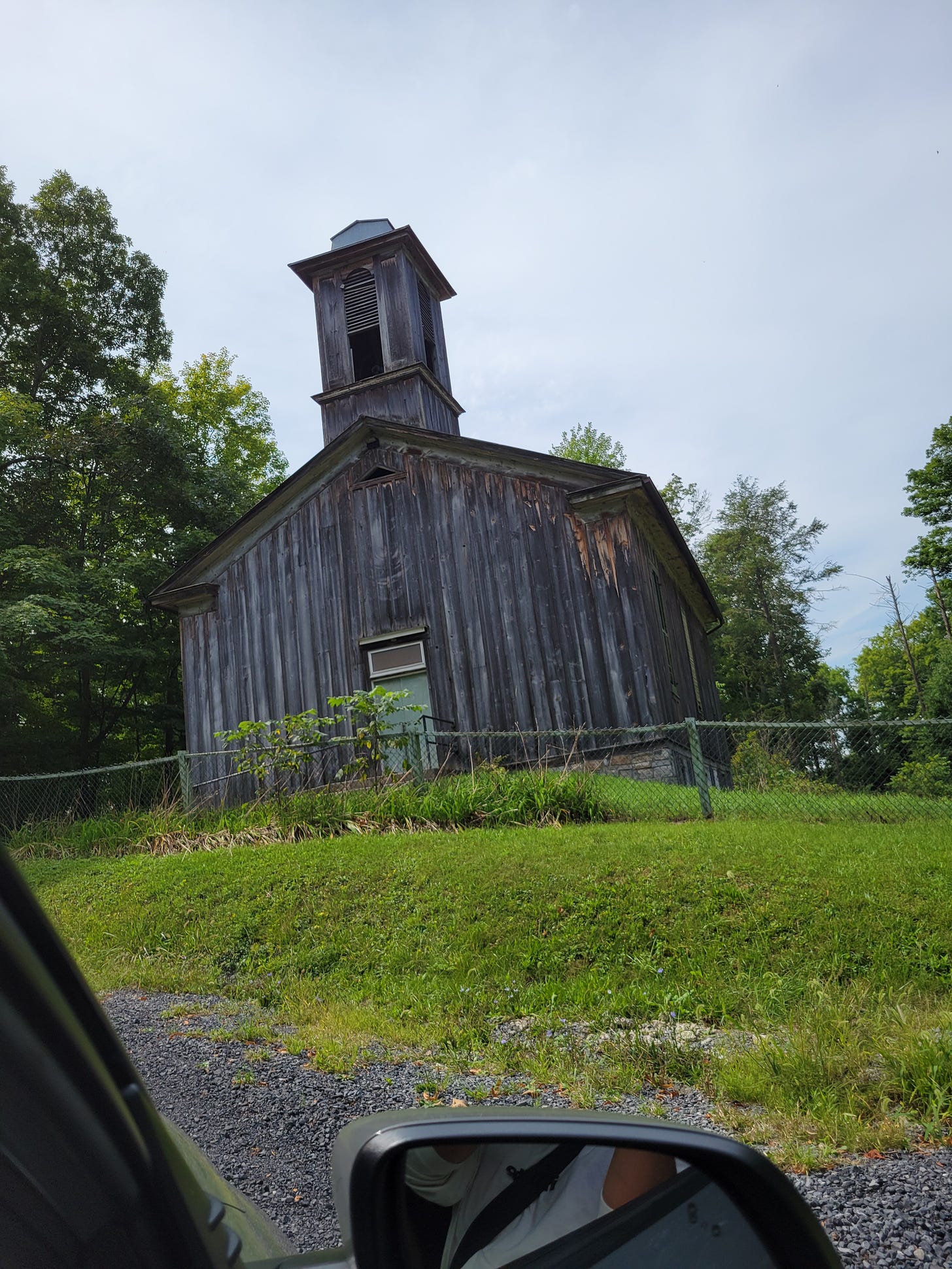 Color photo of a rundown church surrounded by green trees and grass.
