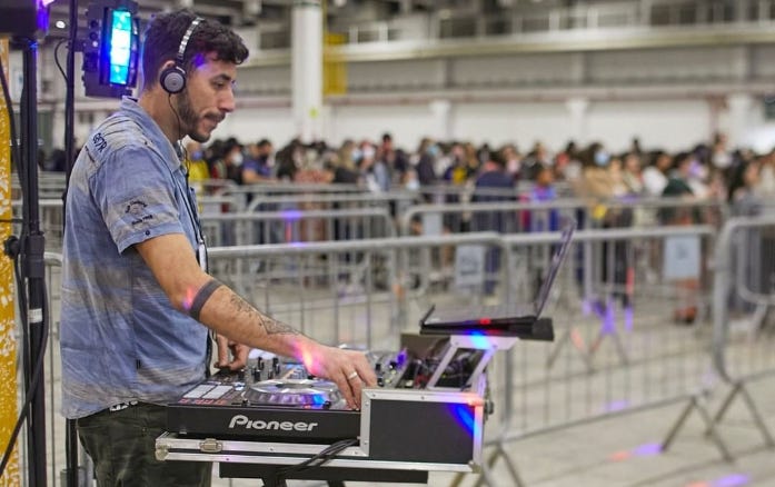 Em primeiro plano, um homem branco com fones de ouvido toca em um equipamento de DJ. Ao fundo, cercadas por grades, muitas pessoas se organizam em uma longa fila.