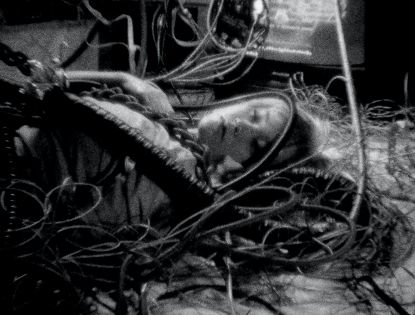 A person wrapped up in wires on the floor. A TV is on in the corner of the frame.