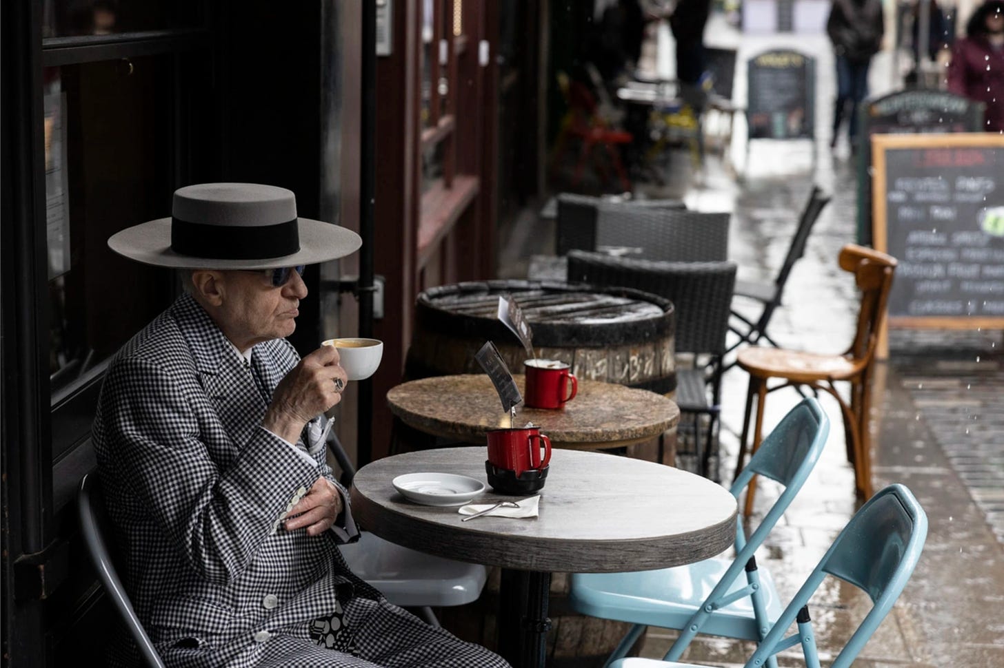 An older gentleman in a stylish suit and broad brimmed hat sipping a latte alone at an ourdoor cafe table under an awning in the London rain, looking pretty grumpy, to be honest.