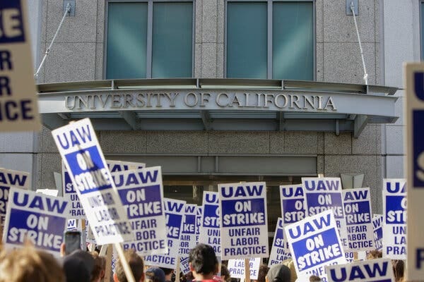 The heads of protesters are visible alongside their signs declaring “U.A.W. on strike — unfair labor practice.” They are standing beneath a University of California sign outside a building.