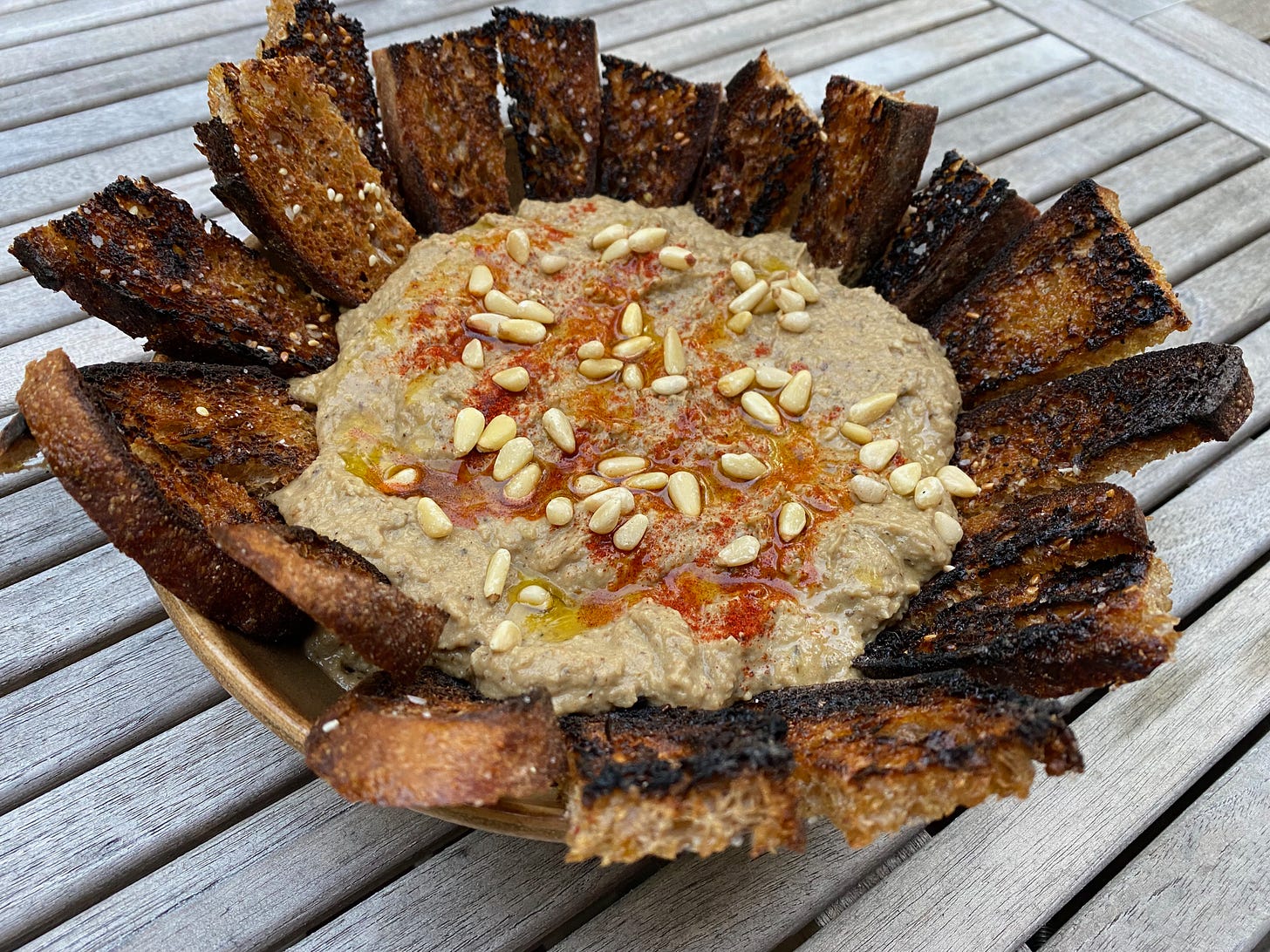 A bowl of baba ganoush topped with pine nuts and paprika, with toasty slices of bread arranged in a ring around the edges.