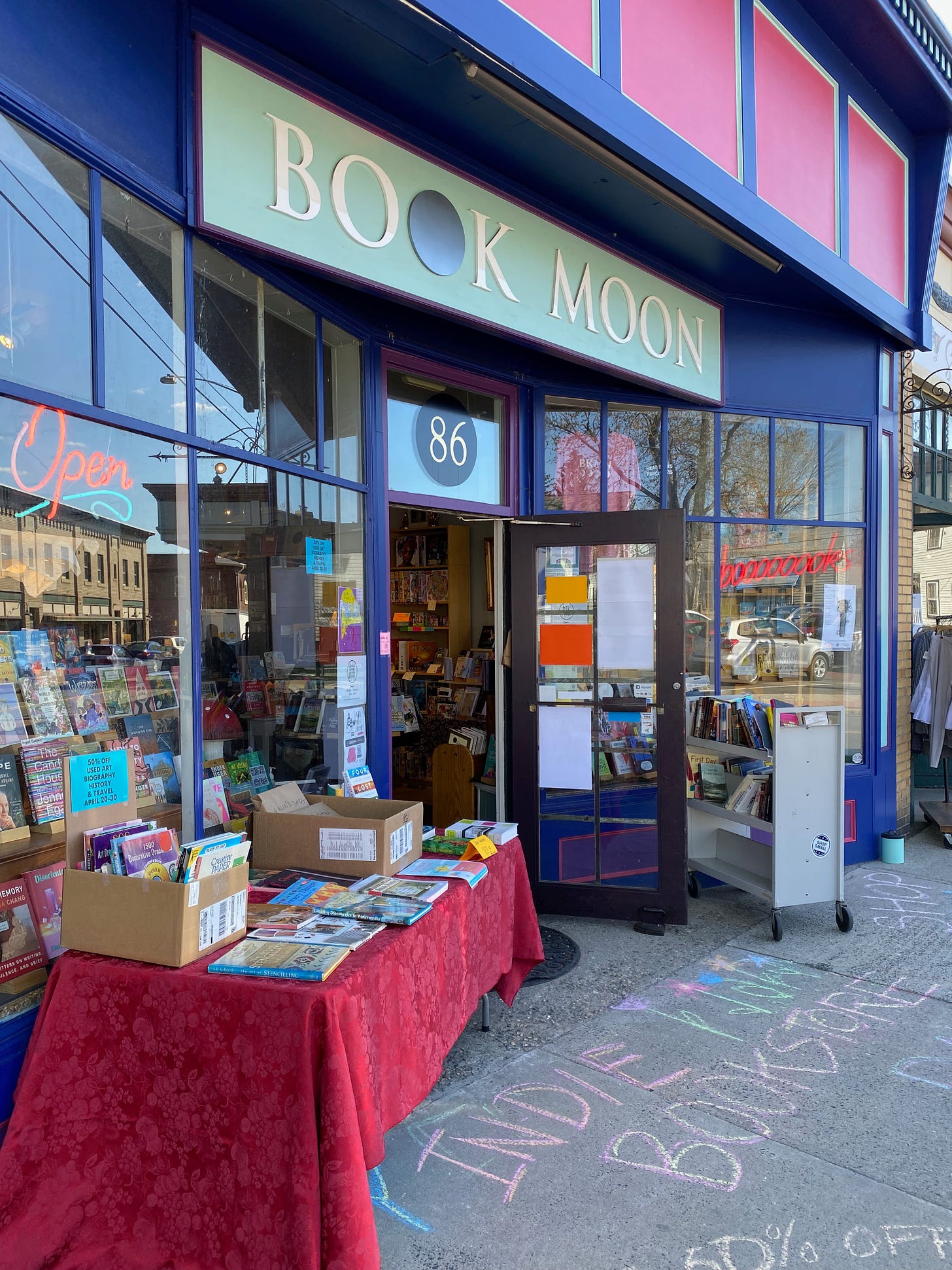 The entrance to Book Moon Books, a blue and pink storefront with several tables and carts of books out front, and many more displayed just inside the open door. Indie Bookstore Day is chalked in bright colors on the sidewalk.