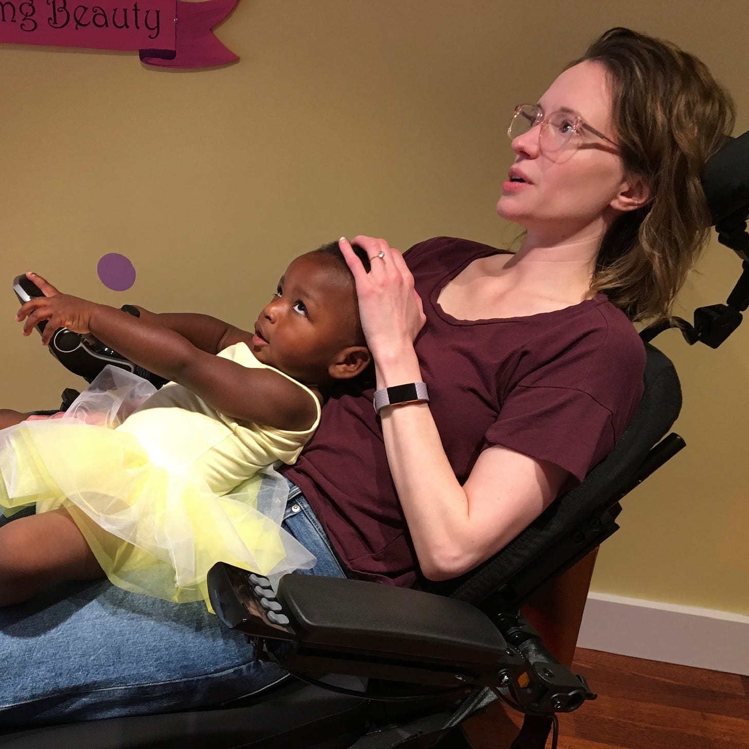 A Black child on the lap of a white woman in a wheelchair. The child is wearing a yellow tutu. The woman is wearing a red shirt. Both are kind of rolling their eyes. 