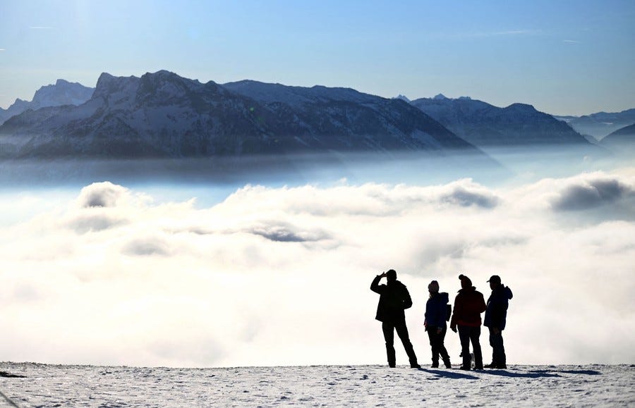 Four people stand on a mountain, looking out over a cloud-filled alpine valley.