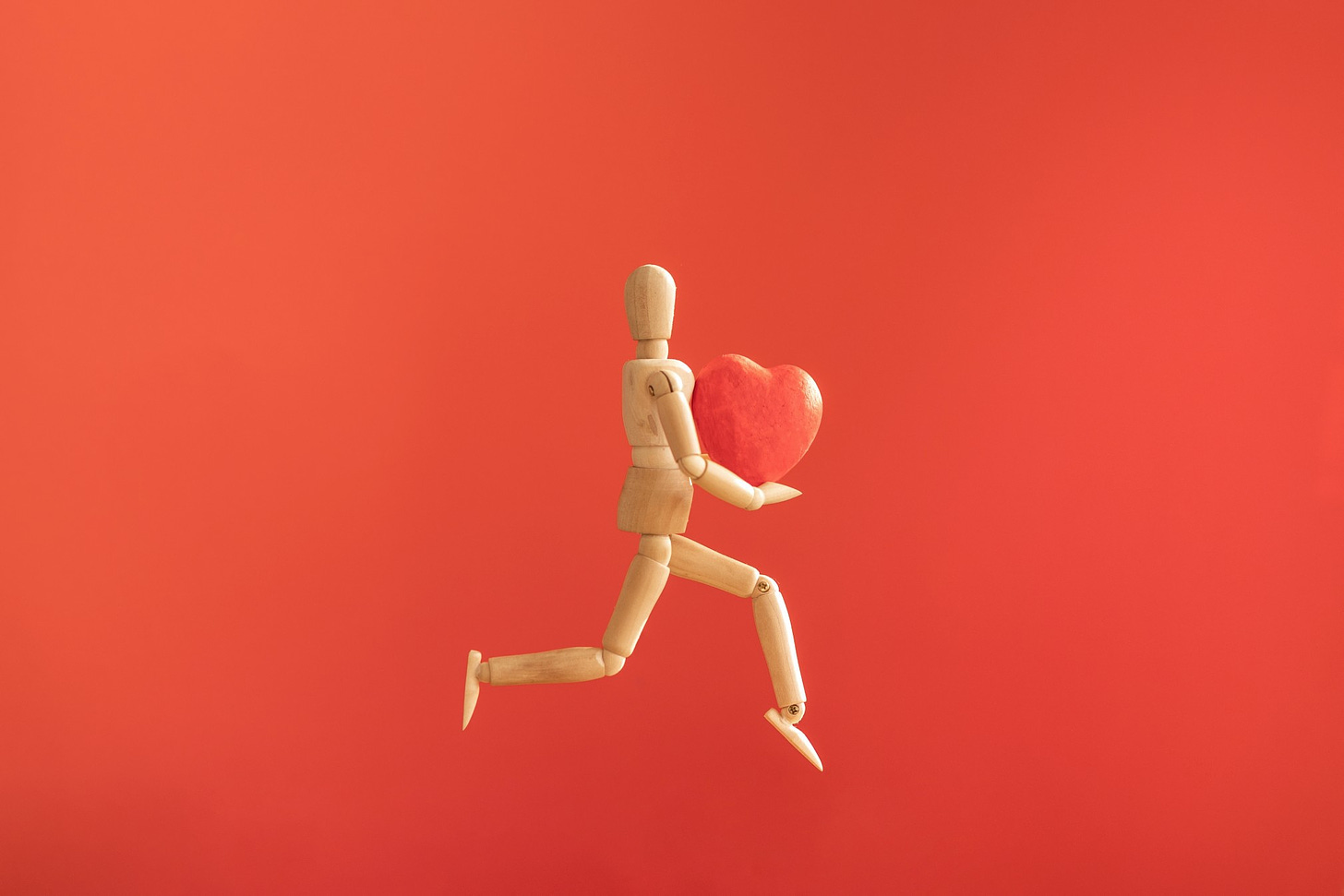 A wooden modeling figure poses in a jumping motion while holding a large heart (against a red background).