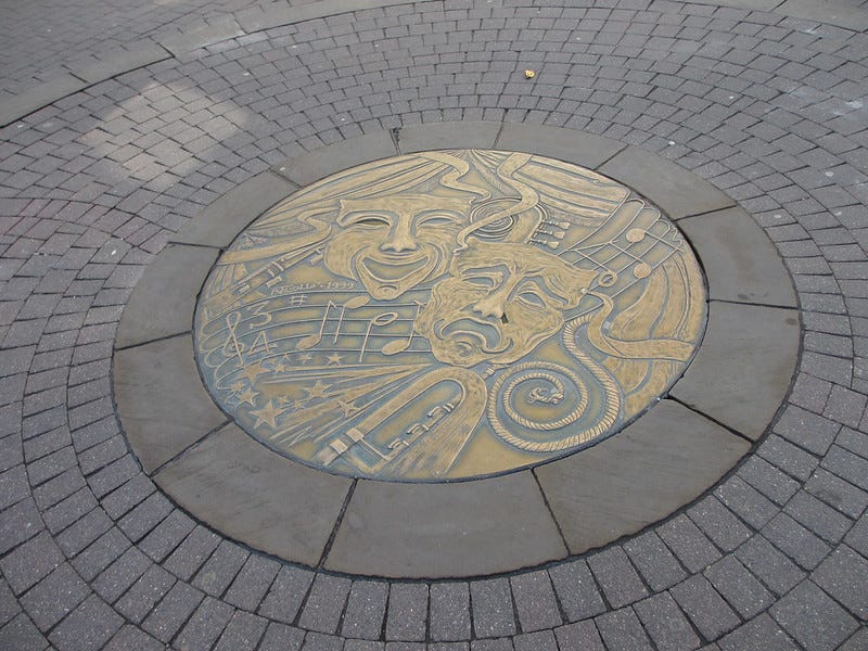 A photograph of pavement stones with a round metal inlay depicting tragedy and comedy masks and musical imagery.