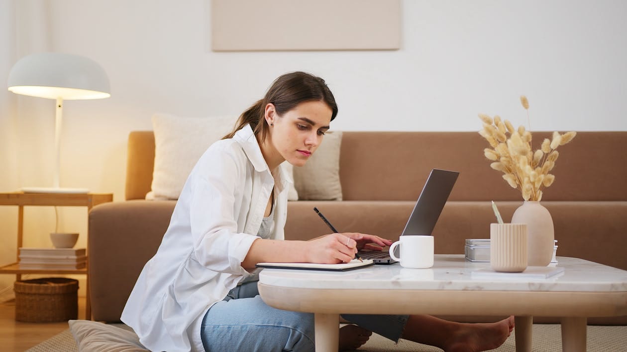 Side view full length young focused female in casual clothes browsing modern laptop and writing down notes while sitting on floor in cozy modern apartment