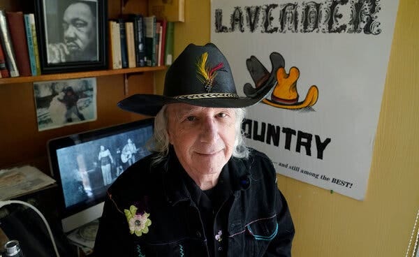 Patrick Haggerty, in a black cowboy shirt and hat, in front of bookshelf and a wall poster that says “Lavender Country.”