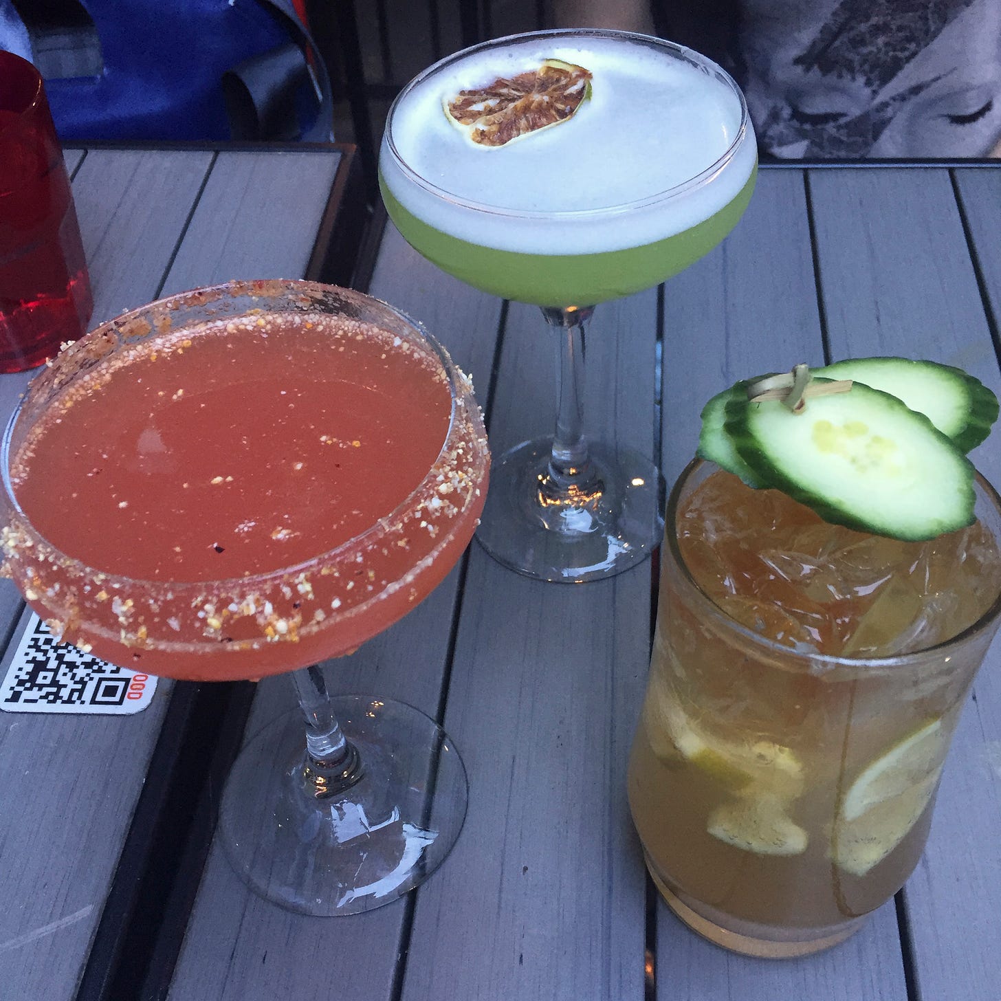 Three drinks on a patio table: one pink in a martini glass, one lime green in a coupé glass with a dehydrated lime floating in the foam on top, and one in a collins glass full of ice with cucumber slices speared on a toothpick.