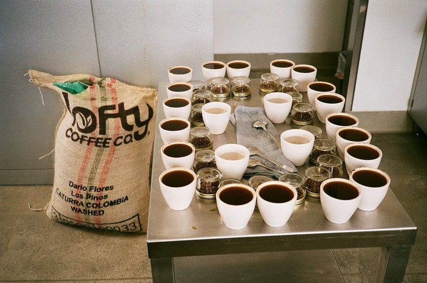On the left a burlap coffee bag full with the Lofty Coffee Logo. Center and right is a stainless steel rolling table with coffee cupping bowls full of coffee, beans and cupping spoons around the edge filling the entire table.