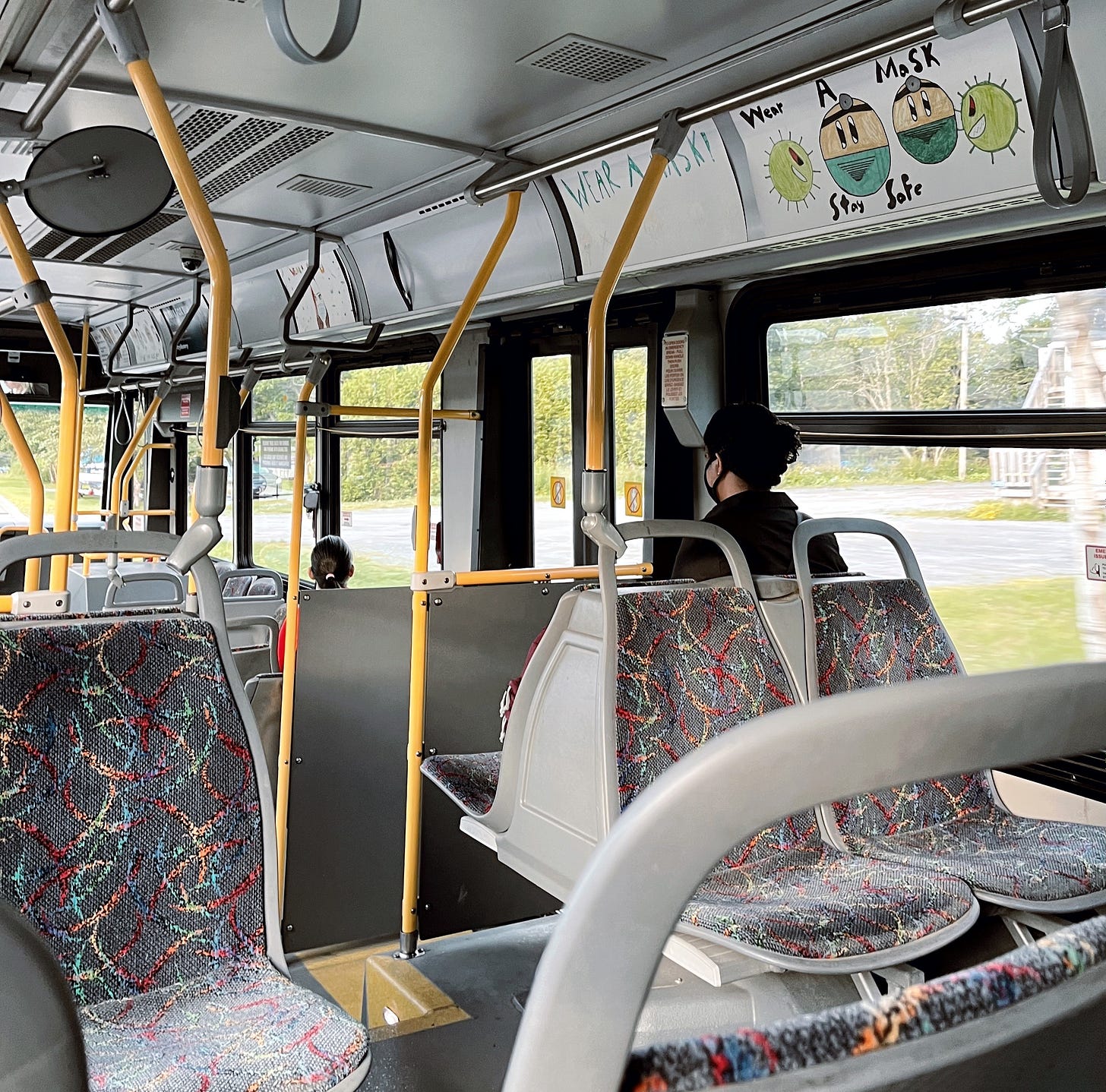 Interior of Saint John Transit bus. Focus is on one of the advertising panels, which is a hand-drawn image by a child, of two yellow face emojis wearing masks, flanked by sneering coronavirus particles. The message reads, "Wear a Mask. Stay Safe"