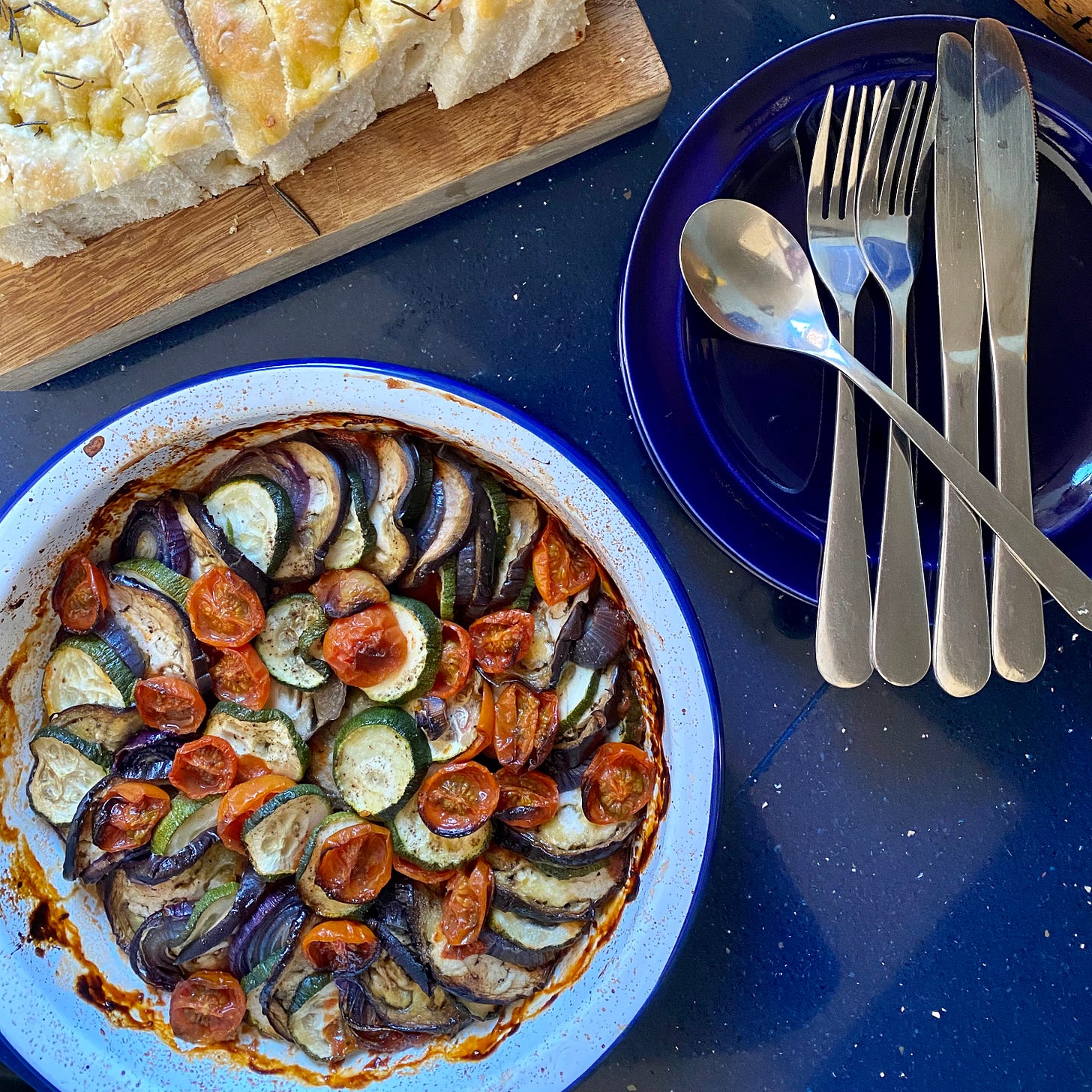 Dish of ratatouille, with a chopping board with slices of focaccia above, and plates and cutlery to the right. 